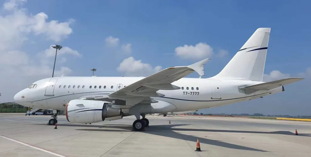A white Airbus A319-style private jet with registration T7-7777 parked on an airfield under a blue sky.