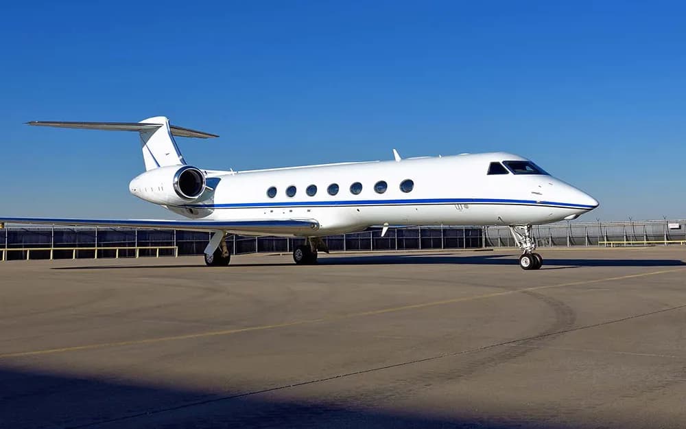 A white Gulfstream G550 business jet with a blue stripe parked on the tarmac under a clear blue sky.