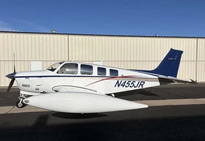 A white Beechcraft Bonanza N455JR with blue and red stripes is parked on an airfield next to a hangar.