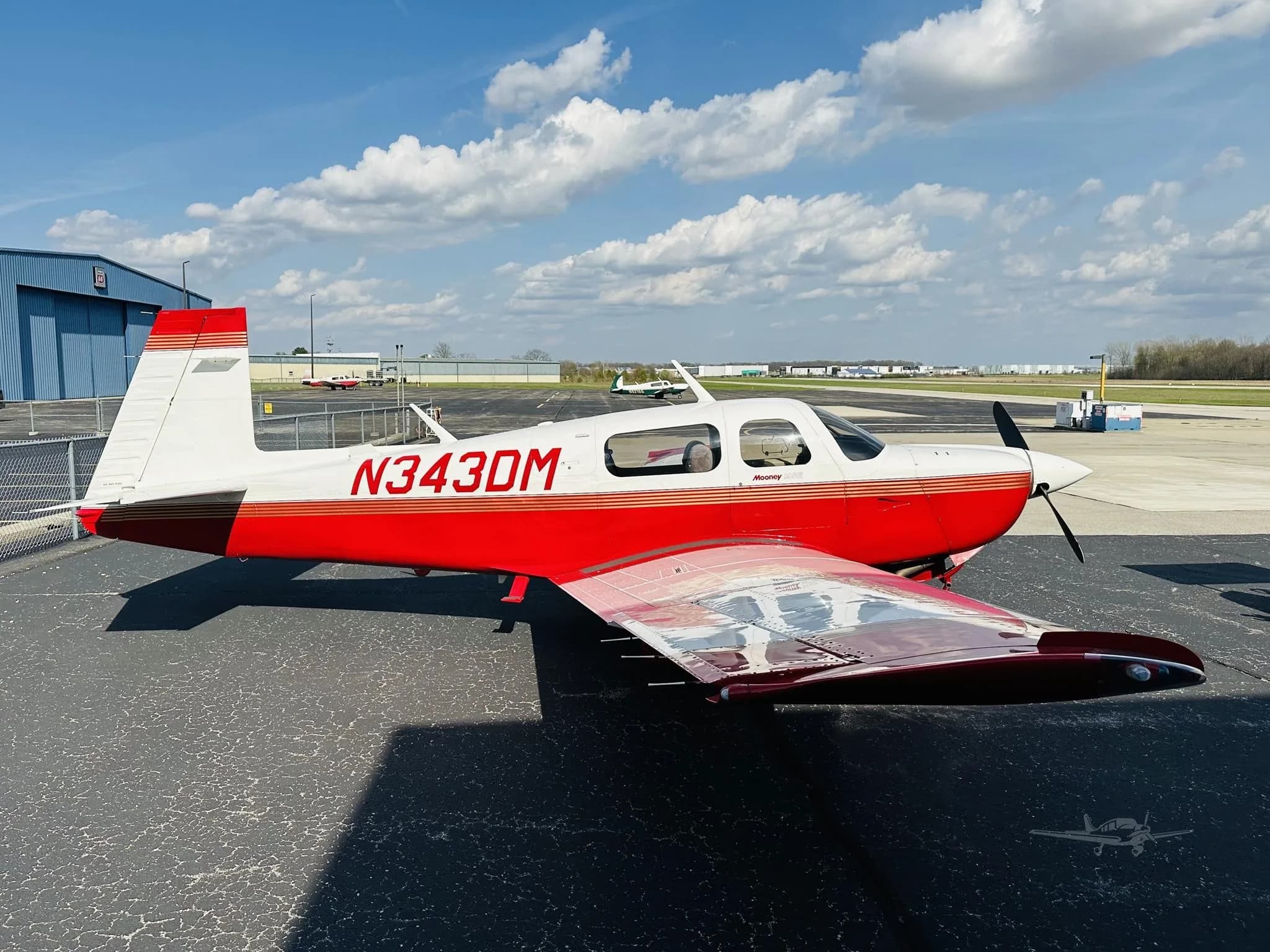 A red and white Mooney M20 aircraft with registration N343DM parked on an asphalt tarmac under a partly cloudy sky.