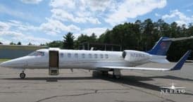A white Learjet business jet with blue tail accents is parked on an airfield with its main door open.