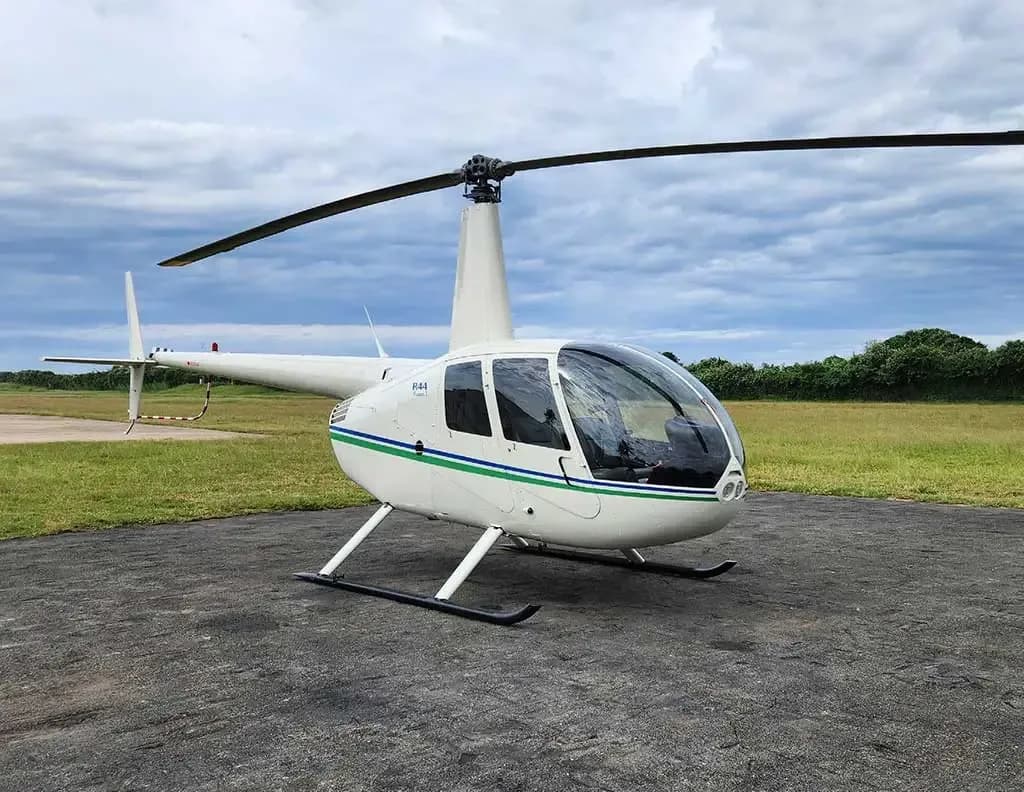 A white Robinson R44 helicopter with green and blue stripes on its fuselage, parked on an asphalt pad under a cloudy sky.