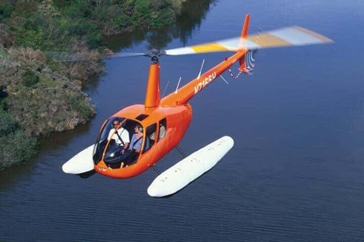 An orange Robinson R44 helicopter with floats, registration N715BU, flying low over a river with two people inside.