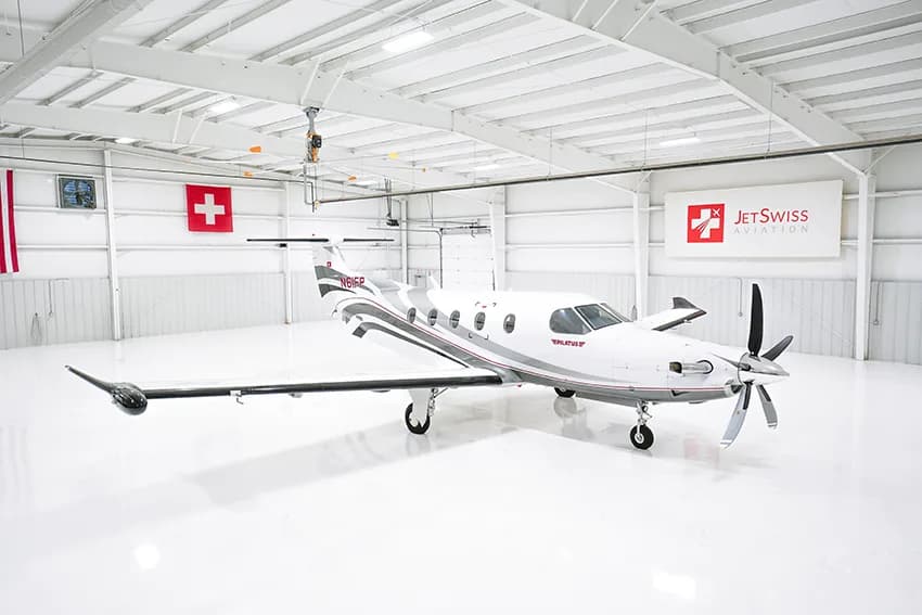 A white Pilatus PC-12 aircraft with gray and red stripes is parked in a brightly lit hangar.