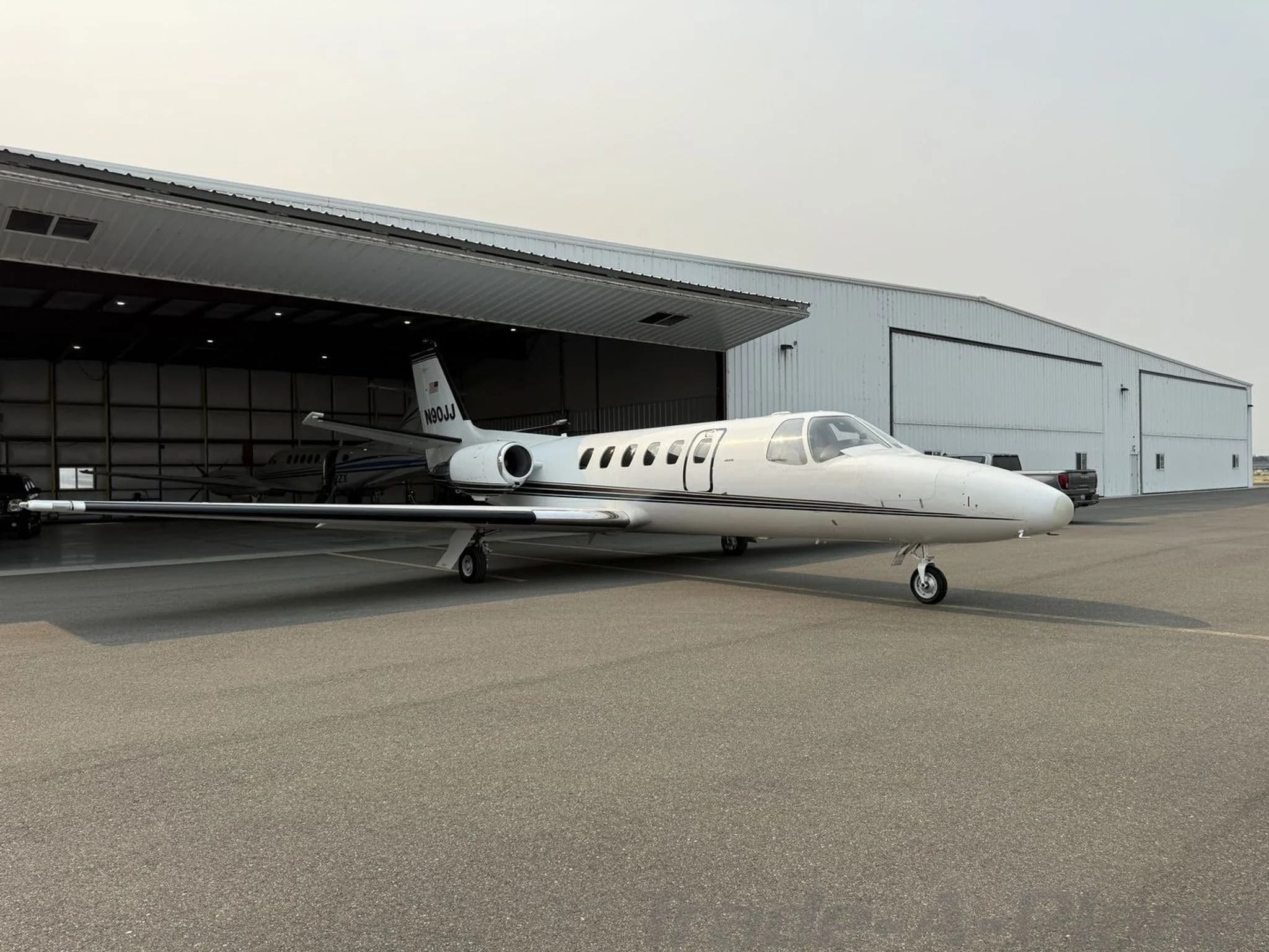 A white Cessna Citation jet, N90JJ, with black and gold stripes, parked on an tarmac in front of an open hangar.