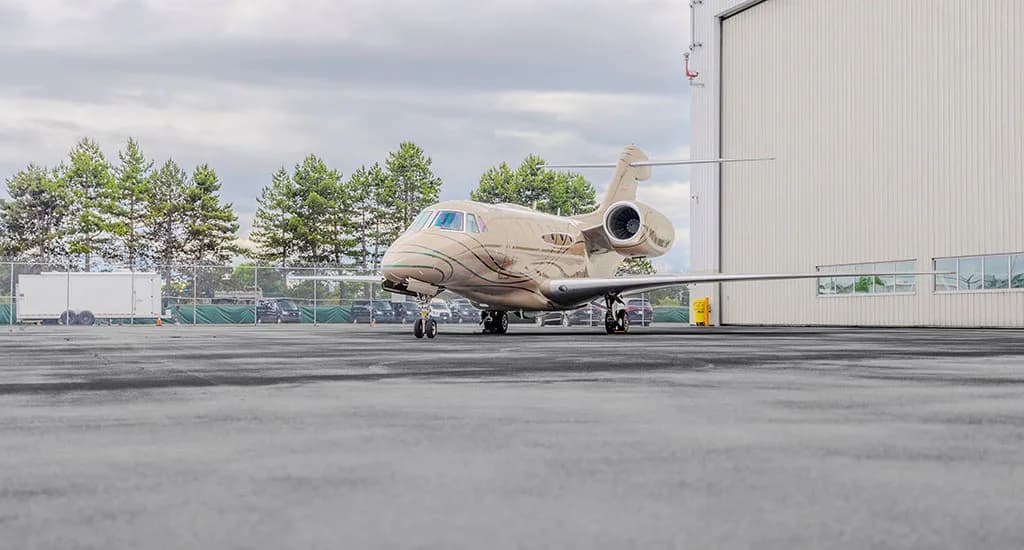 A beige business jet with green and brown stripes parked on an asphalt apron next to a white hangar under a cloudy sky.