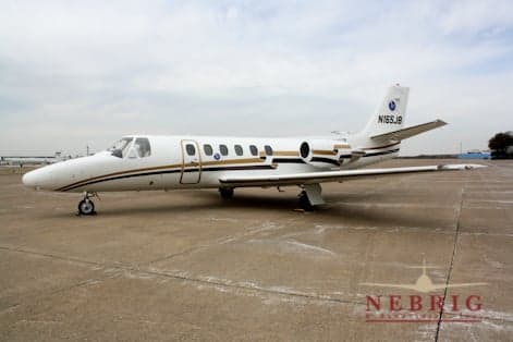 A white Cessna Citation business jet with gold and black stripes is parked on an airport tarmac under a cloudy sky.