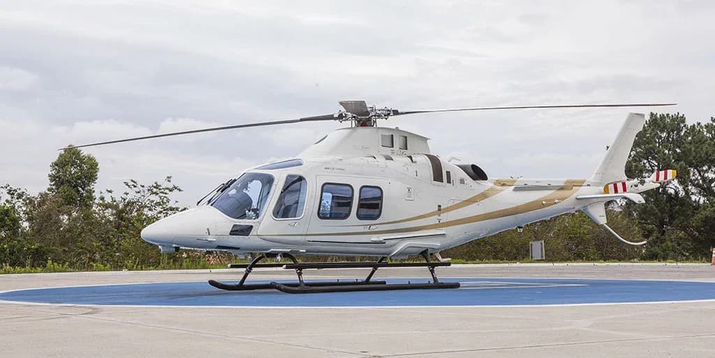 A white helicopter with gold stripes is parked on a blue helipad under a cloudy sky, viewed from the side.