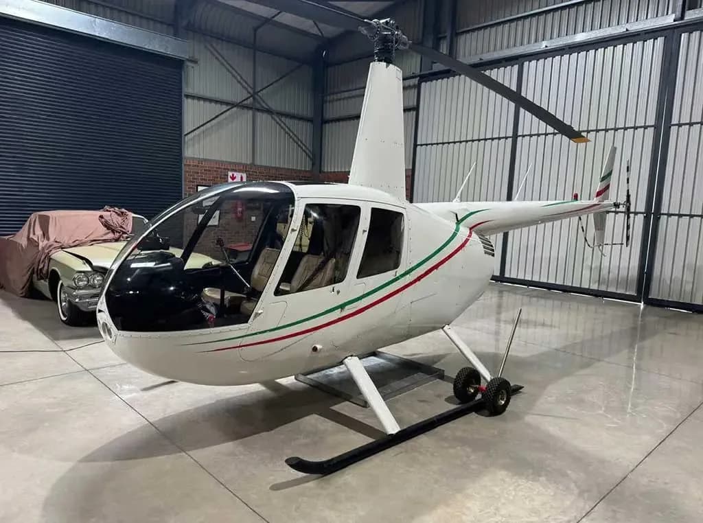 A white Robinson R44 helicopter with green and red stripes is parked inside a hangar, viewed from the front-right.