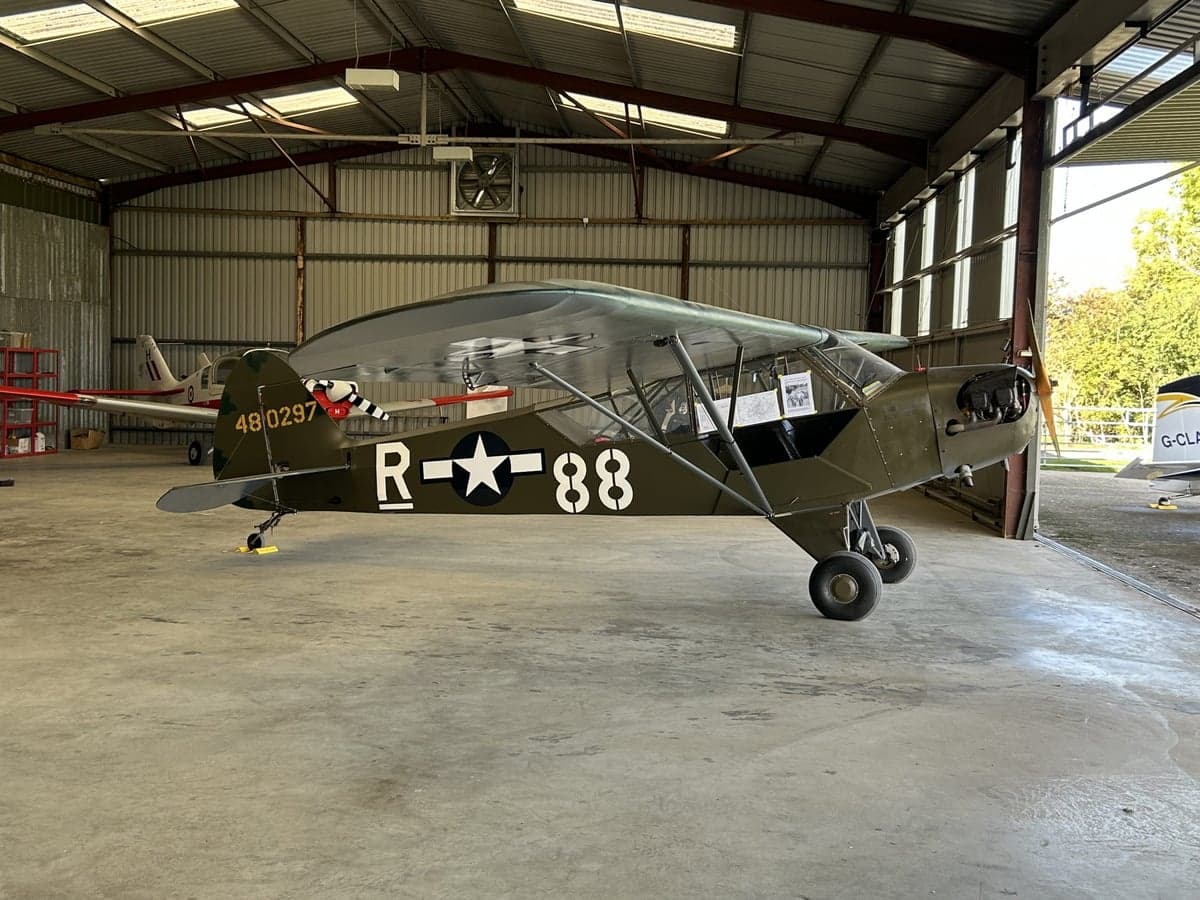A Piper Cub aircraft with military markings parked inside a hangar.