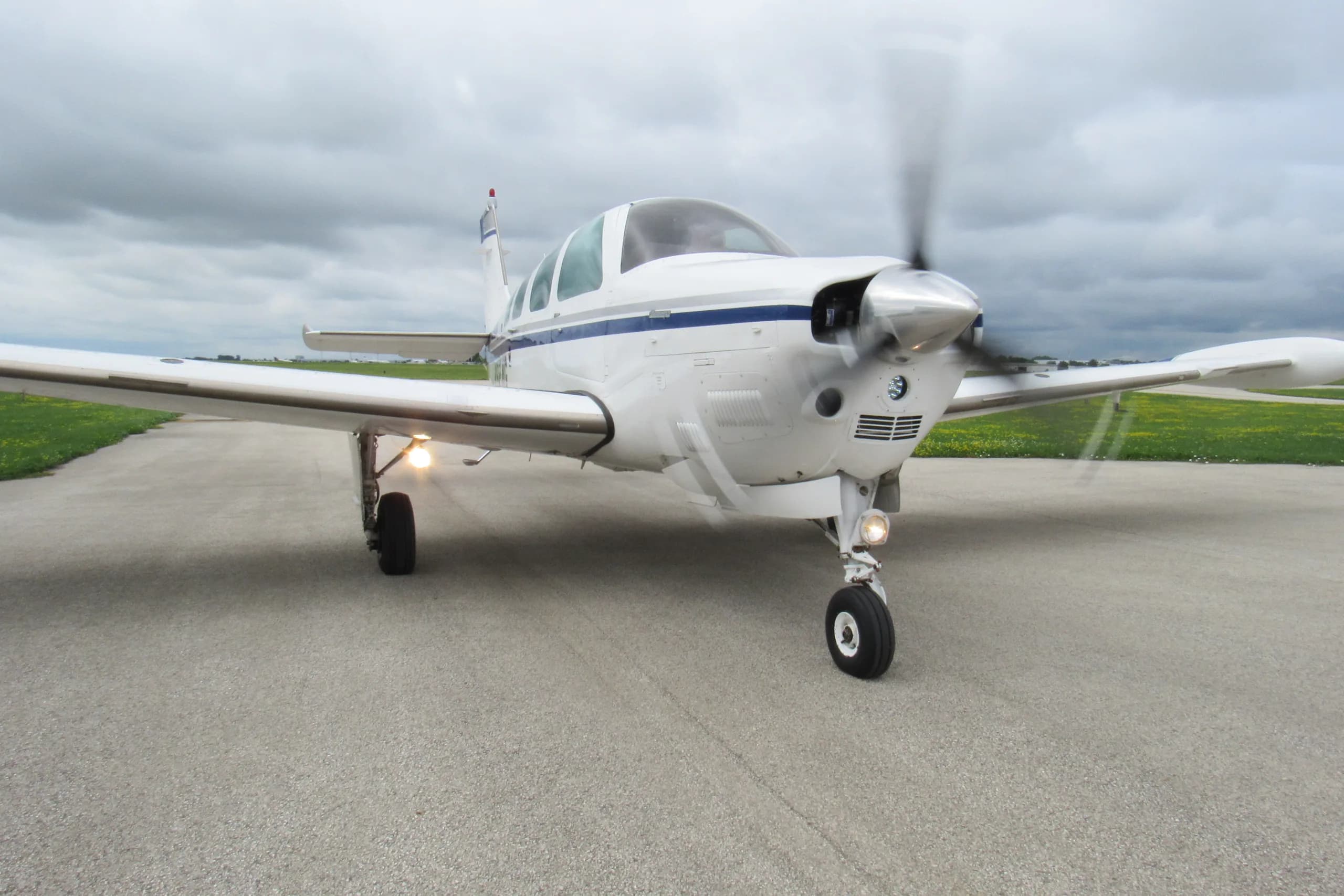 A white single-engine propeller aircraft with blue stripes on a tarmac, with its propeller spinning and landing lights on, under a cloudy sky.
