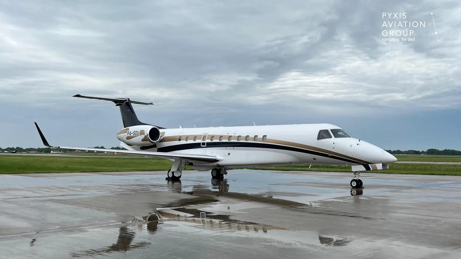 A white and gold Embraer Legacy 600 business jet, registration P4-SDU, parked on a wet tarmac under a cloudy sky.