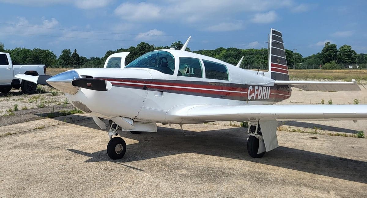 A white Mooney M20J aircraft with red and grey stripes, registration C-FDRN, parked on an asphalt ramp.