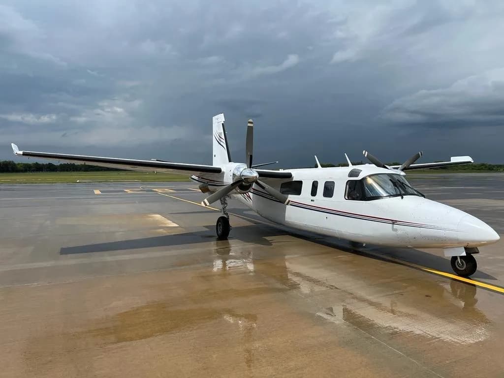 A white twin-engine turboprop aircraft with red and blue stripes is parked on a wet tarmac under a dramatic cloudy sky.