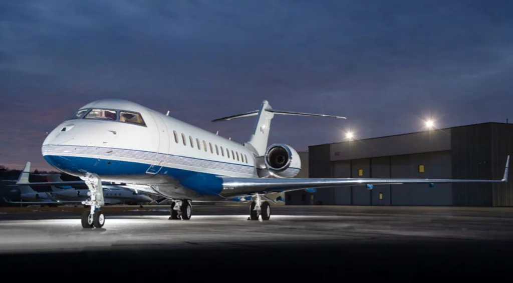 A white and blue business jet parked on an tarmac at dusk with hangar lights in the background.