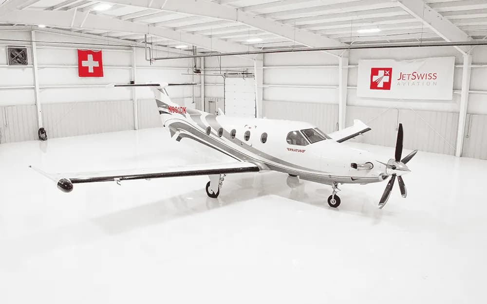A white Pilatus PC-12 aircraft with gray and red accents is parked in a brightly lit hangar.