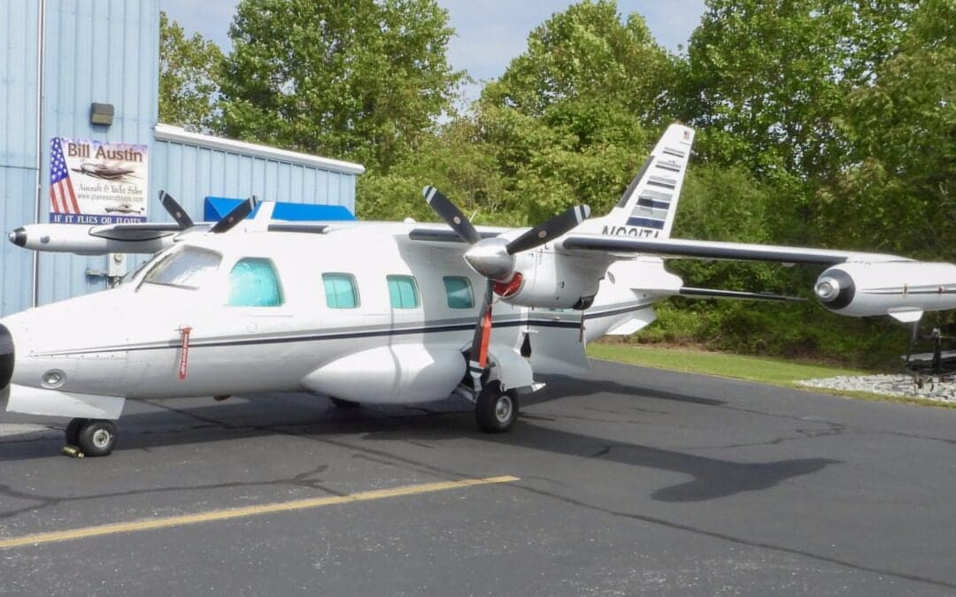 A white Mitsubishi MU-2 aircraft with black and grey stripes on the tail, parked on an asphalt ramp next to a blue building.