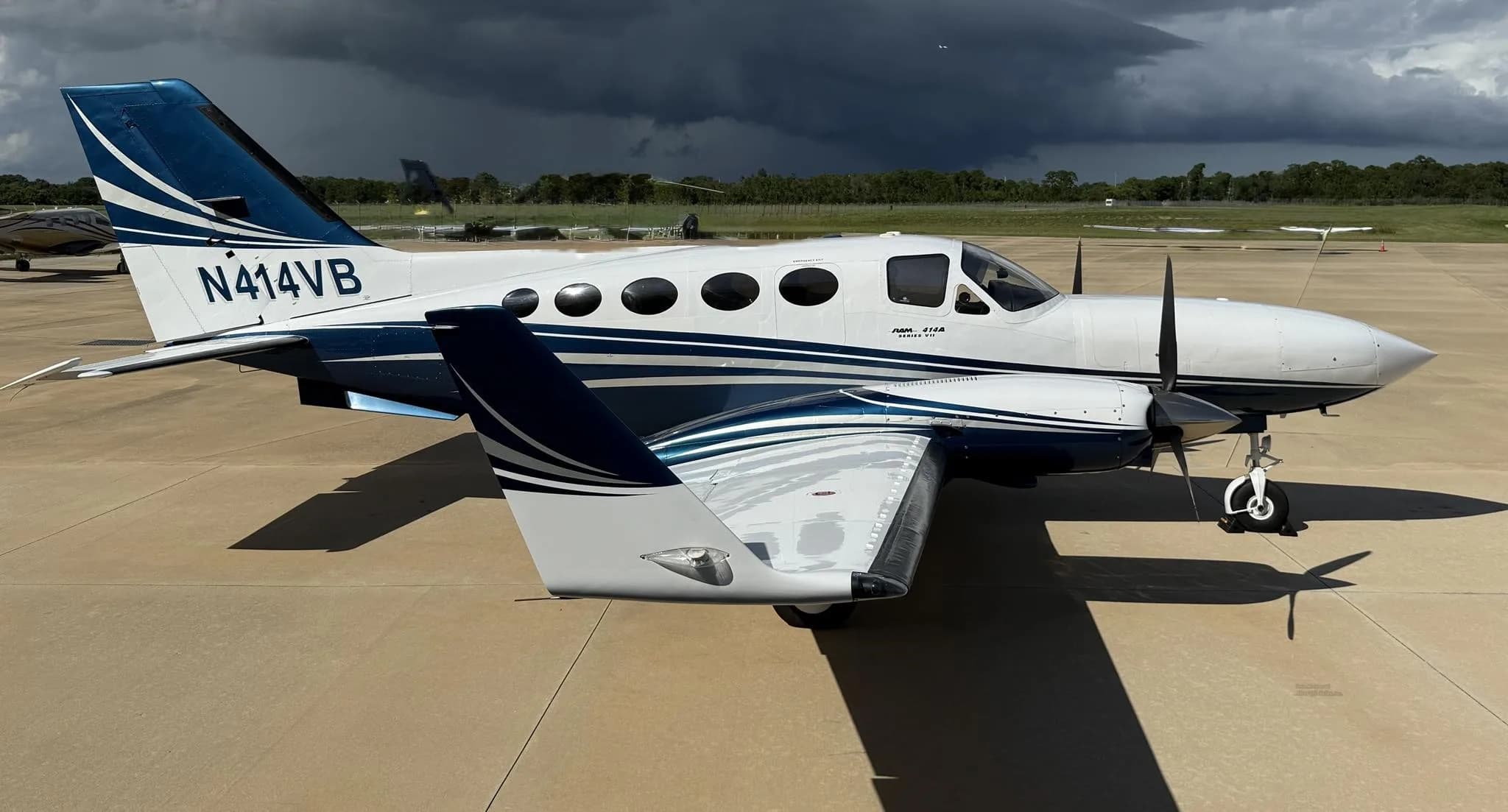 A white and blue Cessna 414 Chancellor twin-engine aircraft with tail number N414VB parked on an airfield under a cloudy sky.