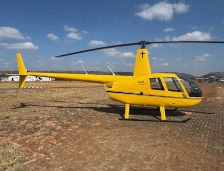 A bright yellow Robinson R44 helicopter with a black stripe and a cross symbol on its tail fin, parked on a dirt airfield under a blue sky.