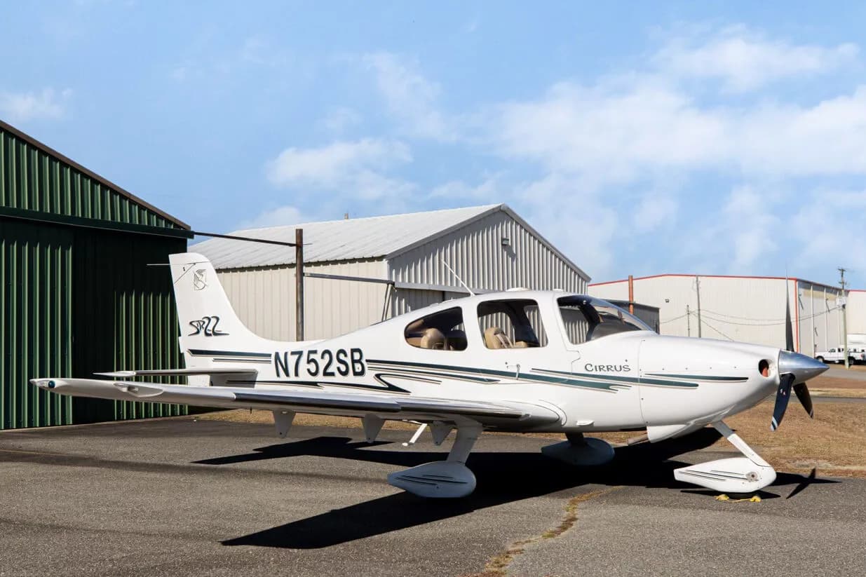 A white Cirrus SR22 aircraft with green and grey stripes, registration N752SB, parked on an asphalt ramp next to a green hangar.