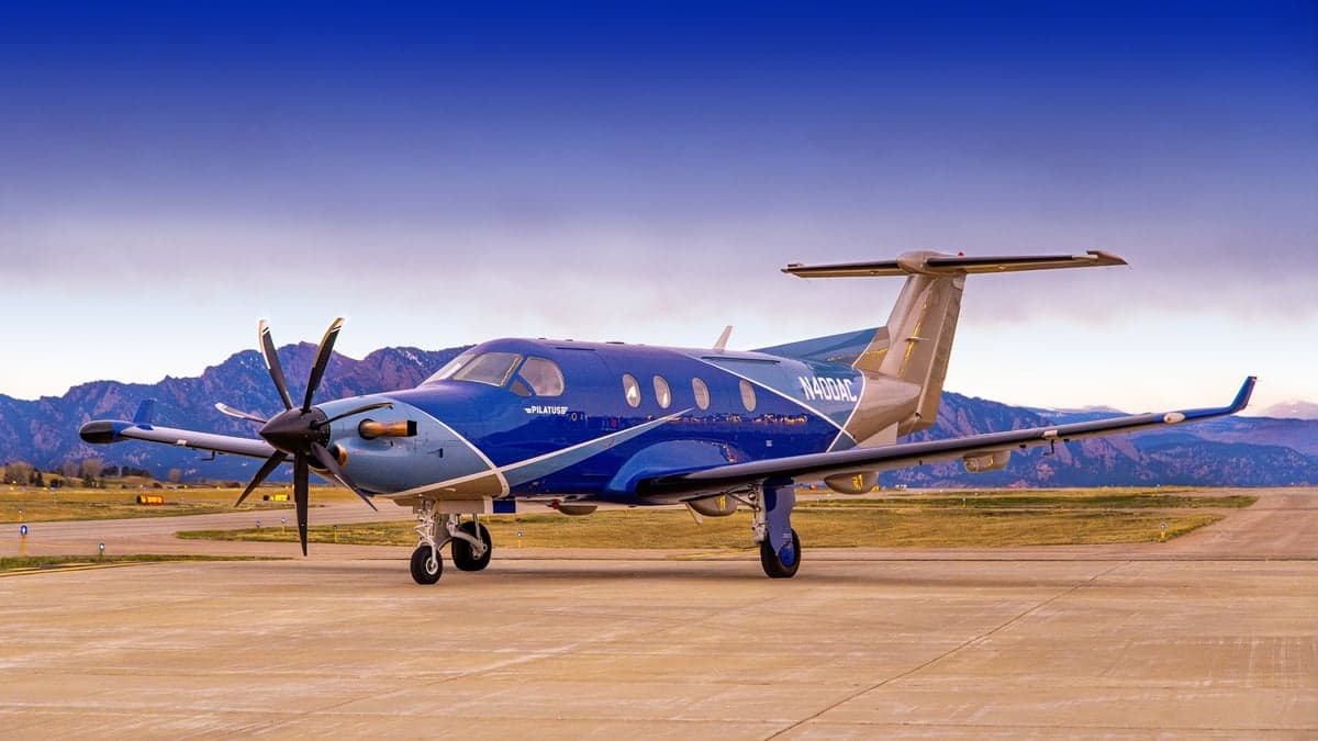 Pilatus PC-12 aircraft with a blue and silver paint scheme parked on a runway with mountains in the background.