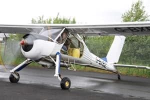 A white and blue Rans S-6ES Coyote II light aircraft with a black spinner is parked on wet tarmac.