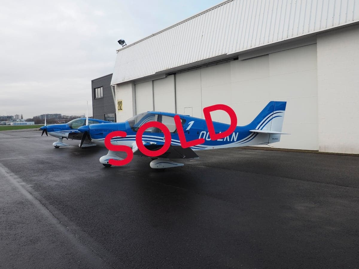 A blue and white single-engine aircraft, registration OO-LKN, parked on a wet tarmac next to a hangar with 'SOLD' text overlaid.
