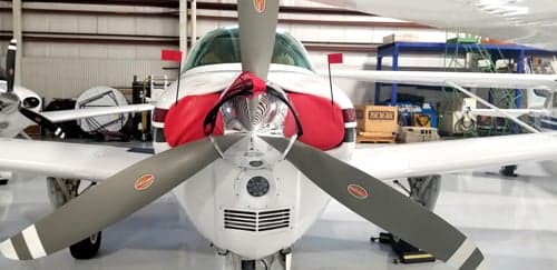 A front view of a white aircraft with a red engine cover and a three-bladed propeller, parked in a hangar.