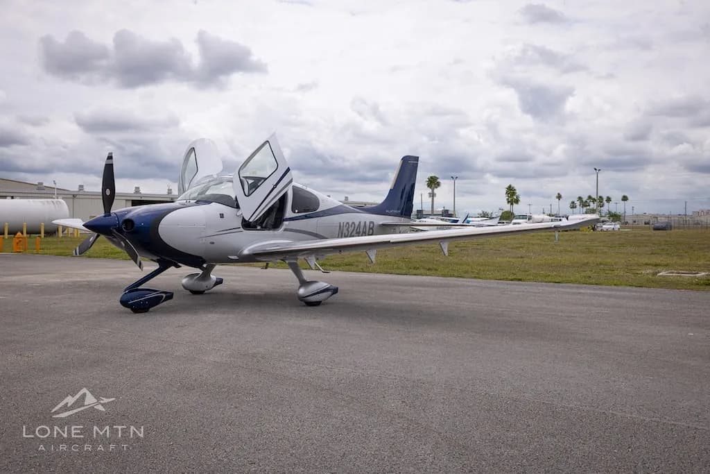 A white and blue Cirrus SR22T aircraft with open doors parked on an airfield, viewed from the front-left.