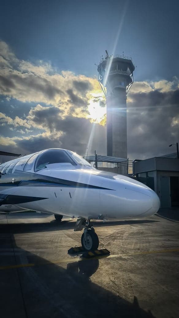 A white Cessna Citation Mustang jet is parked on the tarmac with an airport control tower in the background.