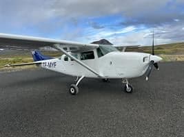 A white Cessna single-engine aircraft with blue tail accents is parked on an asphalt runway under a cloudy sky.