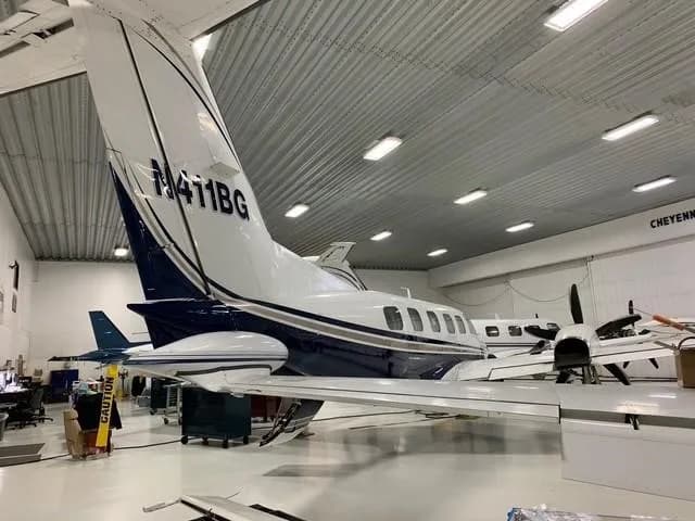 A white and blue twin-engine turboprop aircraft, likely a Beechcraft King Air, is parked inside a hangar, viewed from the rear-right.